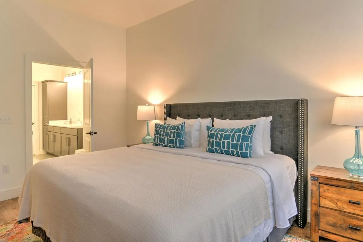A tranquil king bedroom featuring an inviting bed adorned with plush decorative pillows, a classic nightstand, and a warm lamp, all arranged to create a restful, cozy ambiance. The soft lighting and calm decor emphasize the serenity and spaciousness of the retreat.