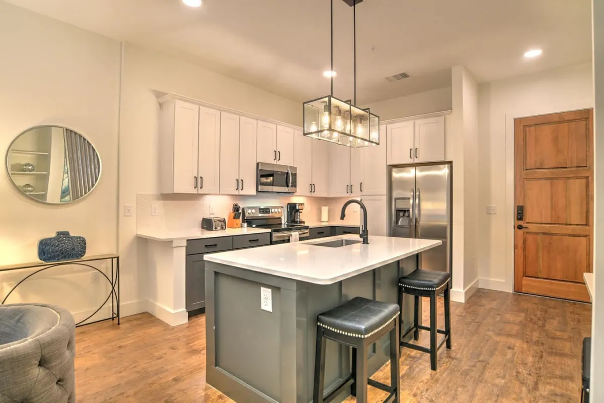 This interior image showcases the condo’s entry area, featuring a stylish wooden door that hints at a secure, contemporary arrival point. The scene includes a modern kitchen with a central island and stainless steel appliances, accentuated by a sleek light fixture. The overall atmosphere is both inviting and secure, blending convenience with comfort.