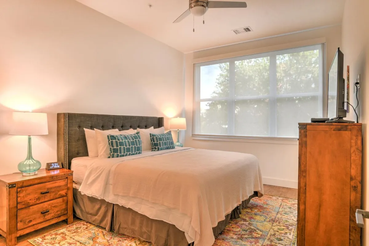 A wide view of the king suite, showcasing the bed, matching nightstands, a large window that lets in abundant natural light, a decorative rug, and a wall-mounted television. The spacious layout hints at both comfort and luxury, with a view toward the outdoors.