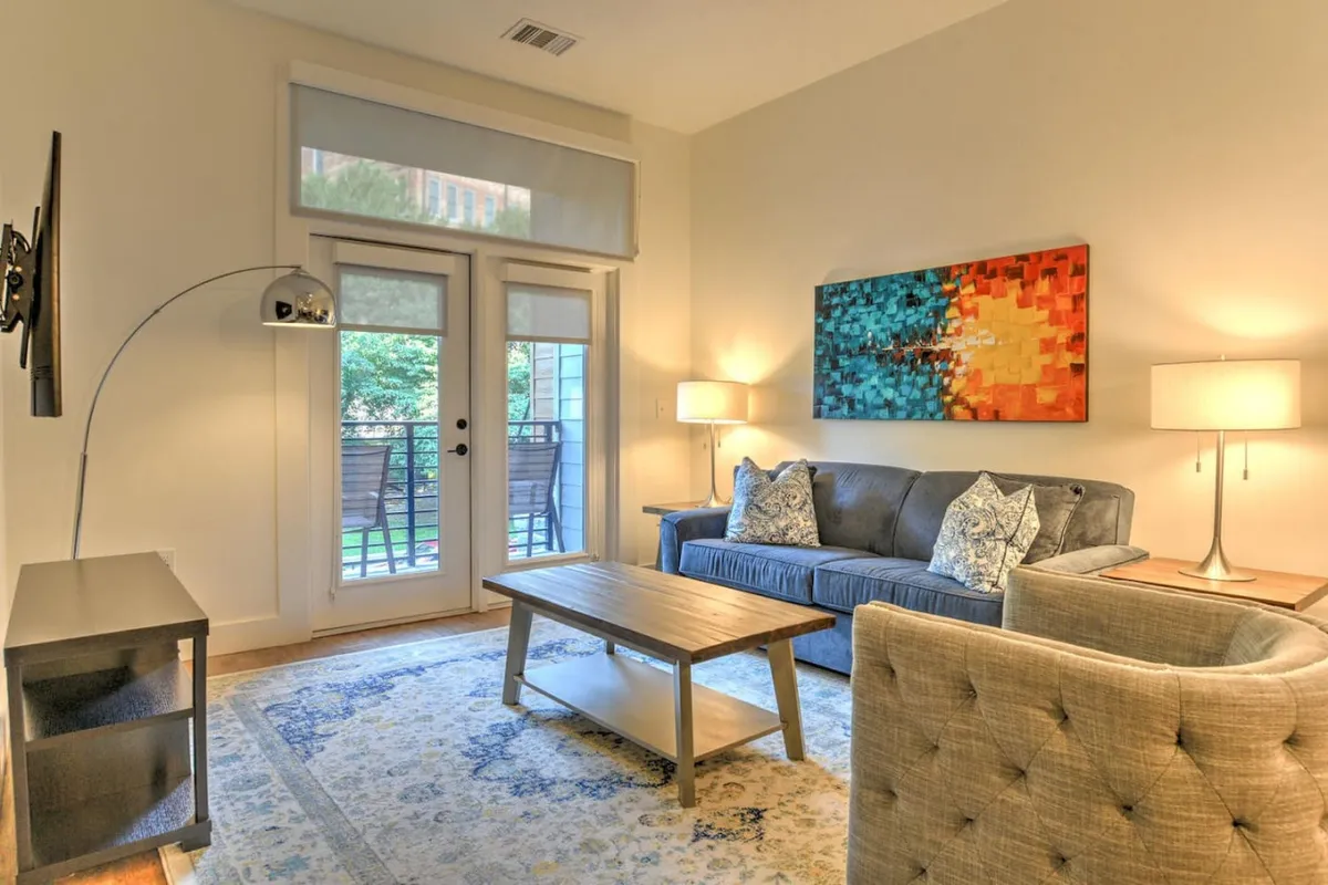 The image showcases the condo's inviting living area with a plush blue sofa, vibrant wall art, a stylish wooden coffee table, ambient floor lamp, and natural light streaming through large windows. The space radiates warmth and comfort, blending classic design with a touch of contemporary flair.