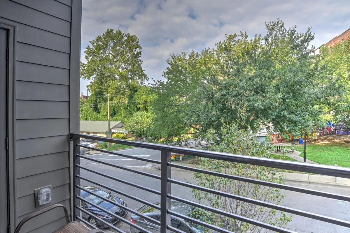 A peaceful view from the balcony, with a railing overlooking lush green trees and a quiet street below. The scene suggests a tranquil spot perfect for enjoying a fresh cup of coffee or stepping out for a breath of morning air, connecting the interior kitchen space to the outdoors.