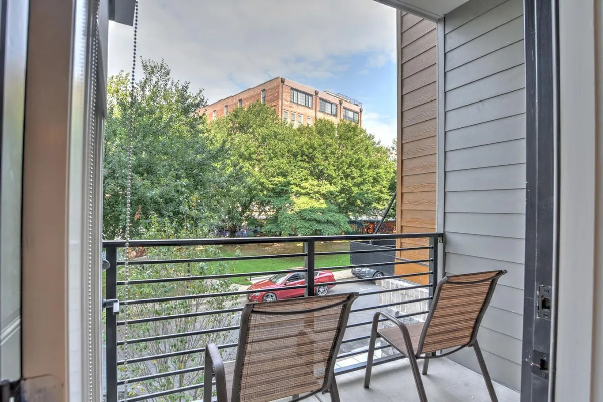 This exterior image highlights the private balcony space with two chairs, lush green trees, and a glimpse of the building across the street. The view connects the parking area and entry to the broader context of urban serenity, suggesting both seclusion and accessibility.